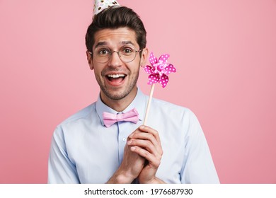 Young White Man Wearing Party Cone Posing With Flower Wind Spinner Isolated Over Pink Background