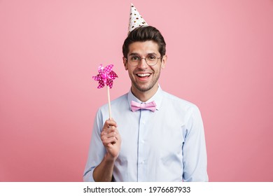 Young White Man Wearing Party Cone Posing With Flower Wind Spinner Isolated Over Pink Background