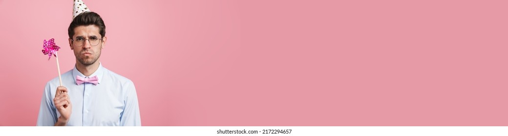 Young white man wearing party cone posing with flower wind spinner isolated over pink background