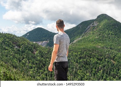 Young white man wearing a t-shirt is staring in the distance contemplating the beautiful view of mountains on a sunny day. Shot on the Ghotics Mountain, Adirondacks National Park, NY, USA. 