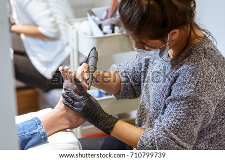 A young white manicurist girl makes a pedicure electric nail drill machine to the client in the workplace. On her face a protective dressing, on her hands protective gloves. Close view.