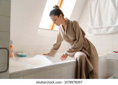 Young white smiling woman in bathrobe bending down over bathtub filled with bubbles, testing water temperature - Powered by Shutterstock