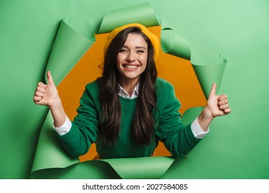 Young white smiling woman peeking out hole isolated on green background thumbs up