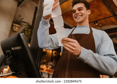 Young white waiter man wearing apron standing in front of computer and holding client receipt in cafe
