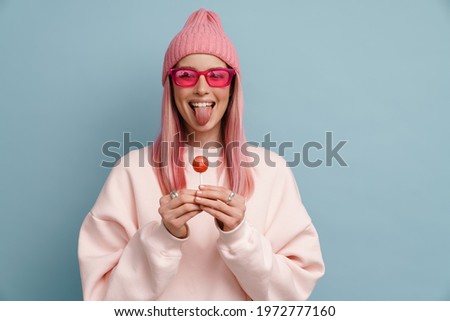Young white woman holding lollipop and sticking out her tongue at camera isolated over blue wall