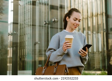 Young white woman smiling while drinking coffee and using cellphone outdoors - Powered by Shutterstock