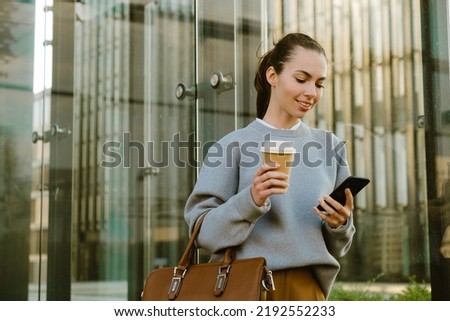 Young white woman smiling while drinking coffee and using cellphone outdoors