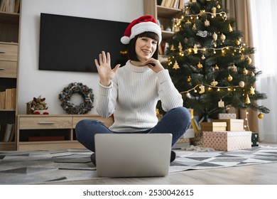 Young white woman wearing Santa hat video chats on laptop at home during Christmas season. She sits on floor in cozy living room with decorated tree and gifts. - Powered by Shutterstock