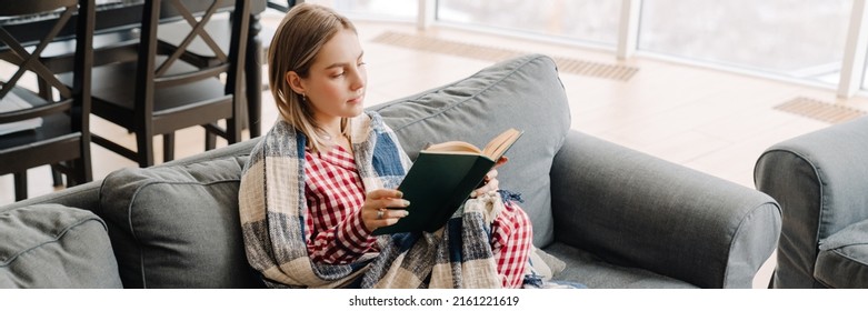 Young white woman wrapped in blanket reading book while sitting on sofa at home