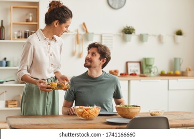 Young Wife Bringing The Dish And Serving Dinner For Her Husband Who Sitting At The Table In The Kitchen