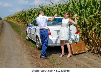 Young wife hitting man with ear of corn behind parked automobile with brown suitcase near cornfield