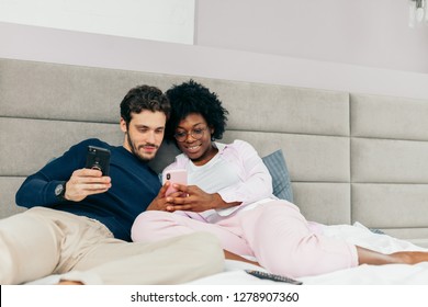 Young wife and husband relaxing on bed during their day-off. Spauses of different nationalities making selfie or watching videos using smart phones