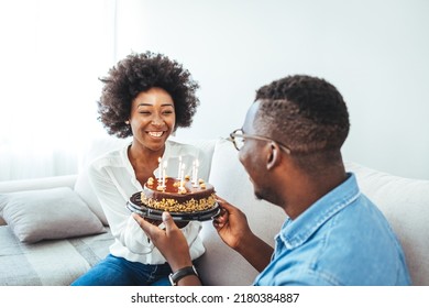 Young Wife Making Surprise For Husband, Excited Man Holding Gift Box And Being So Happy While Sitting In Living Room At Table With Cake. Family Couple In Party Hats Celebrating Birthday At Home