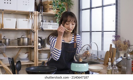 Young Wife Opening Lid And Using Spoon To Try The Delicious Soup On Stove, Smiling With Happiness And Eyes Closed. Background Cozy Kitchen Interior, Real Moments.