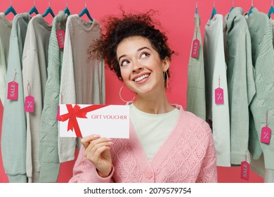 Young wistful female costumer woman wear sweater stand near clothes rack with tag sale in store showroom hold gift certificate coupon voucher card for store isolated on plain pink background studio