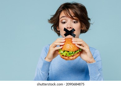 Young Wistful Sad Woman With Mouth Sealed With Tape Wearing Sweater Holding Burger Want To Eat Isolated On Plain Pastel Light Blue Background Studio Portrait. People Lifestyle Diet Junk Food Concept