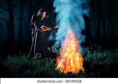 A young witch reading conjure from book at the fire at night in the forest. Priestess of fire. Fantasy illustration. Fairy tales - Shutterstock ID 681782428