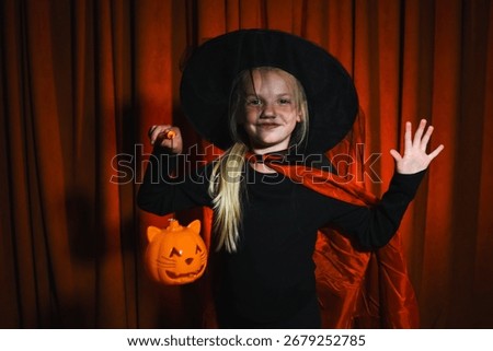 Young witch smiles happily while holding a pumpkin lantern under a warm orange backdrop during a festive Halloween celebration