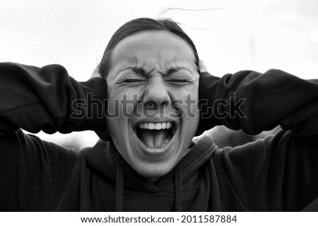 A young woman of 18-25 years old screams with an open mouth, covers her ears with her hands, refuses to listen, closes her eyes, frowns, showing her pain, express emotions, burnout syndrome copy space