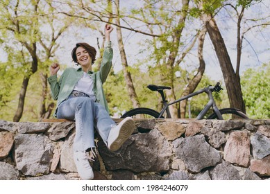 Young woman 20s in casual green jacket jeans sitting on stone bench near bicycle bike in city spring park outdoors do winner gesture clench fist. People active urban healthy lifestyle cycling concept - Shutterstock ID 1984246079