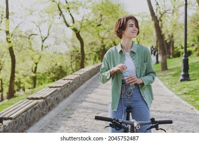 Young woman 20s in green jacket jeans riding bicycle bike in city spring park outdoors, look aside drink clear fresh pure water from transparent glass of water People active healthy lifestyle concept. - Shutterstock ID 2045752487