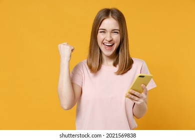 Young Woman 20s Wearing Casual Basic Pastel Pink T-shirt Holding Mobile Cell Phone Surfing Browsing Internet Do Winner Gesture Clench Fist Celebrating Isolated On Yellow Background Studio Portrait.