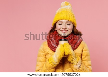 Young woman 20s wears yellow jacket hat mittens keep eyes closed hold takeaway delivery craft paper brown cup coffee to go smell sniff isolated on plain pastel light pink background studio portrait