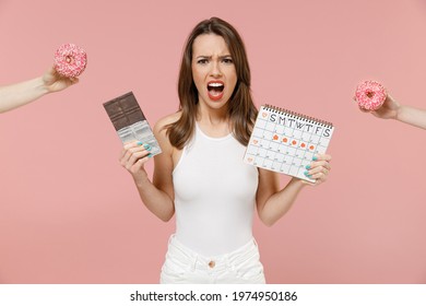 Young woman 20s in white clothes hold female periods calendar check menstruation day bar chocolate hand give donuts scream isolated on pastel pink background Medical healthcare gynecological concept