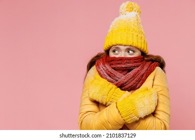 Mujer joven de 20 años usa un sombrero de chaqueta amarilla que imita y mira de lado la cubierta que esconde la boca con la bufanda envuelta en el cuello y las manos cruzadas aislado en un retrato de estudio de fondo rosa claro pastel