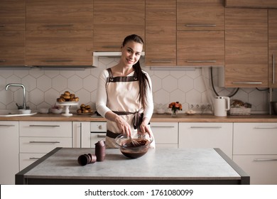 Young woman 25-30 year old cooking homemade chocolate cake at home standing infront of kitchen table mixing batter in bowl closeup. Looking at camera. 20s. 