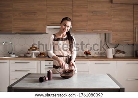 Young woman 25-30 year old cooking homemade chocolate cake at home standing infront of kitchen table mixing batter in bowl closeup. Looking at camera. 20s. 