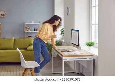 Young woman accountant working from home, standing by her desk, holding a coffee mug, drinking coffee, and looking at financial spreadsheets on her modern desktop computer. Remote work concept - Powered by Shutterstock