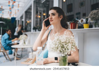 Young woman in activewear sitting at an outdoor cafe, engaged in a phone call. She has a yoga mat and water bottle, surrounded by flowers, enjoying the lively yet peaceful ambiance of the cafe - Powered by Shutterstock