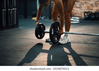 Una joven está agregando pesas a una barra, preparándose para su rutina de ejercicios en un gimnasio. Una fuerte iluminación proyecta una sombra dramática en el piso del gimnasio