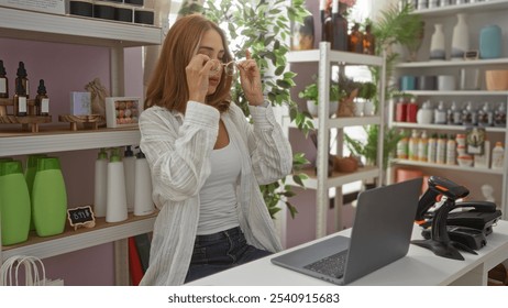 Young woman adjusting glasses in a beautifully decorated home decor store, surrounded by various products on shelves and working at a laptop - Powered by Shutterstock