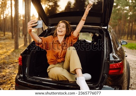 A young woman admires the sunset in the trunk of a car with a phone in her hands. A curly-haired tourist takes pictures on a mobile phone and communicates via video call. Travel, weekend concept.