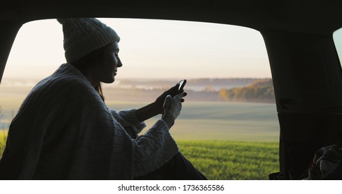 Young Woman Admiring Sunrise In Trunk Of Car Van. Girl Taking Photo On Mobile Phone Against Backdrop Of Nature Panorama In Fog . View From Inside