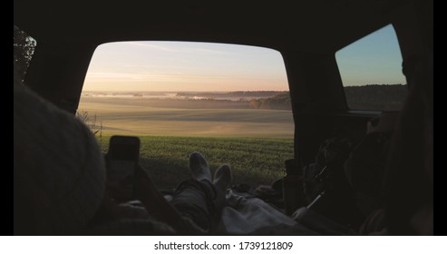 Young Woman Admiring Sunrise In Trunk Of Car Van. Girl Taking Photo On Mobile Phone Against Backdrop Of Nature Panorama In Fog . View From Inside