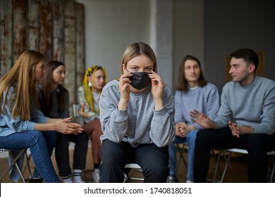 Young Woman Afraid Of COVID-19, She Is Wearing Medical Mask, Sits In The Center And Look At Camera While Group Of People Sit In Background Without Masks. Stay Home