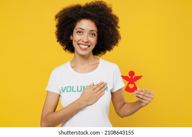 Young woman of African American ethnicity wears white volunteer t-shirt hold in hands little paper angel put hand on heart isolated on plain yellow background. Voluntary free work help grace concept - Shutterstock ID 2108643155