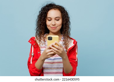 Young Woman Of African American Ethnicity 20s She Wear Red Jacket Hold In Hand Use Mobile Cell Phone In Yellow Case Isolated On Plain Pastel Light Blue Cyan Background. Wet Fall Weather Season Concept