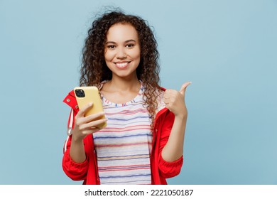 Young Woman Of African American Ethnicity 20s She Wear Red Jacket Hold In Hand Use Mobile Cell Phone Show Thumb Up Isolated On Plain Pastel Light Blue Cyan Background. Wet Fall Weather Season Concept