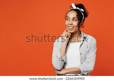 Young woman of African American ethnicity she wears grey shirt headband put hand prop up on chin, lost in thought and conjectures isolated on plain orange background studio. People lifestyle concept