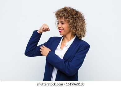 young woman african american feeling happy, satisfied and powerful, flexing fit and muscular biceps, looking strong after the gym against flat wall