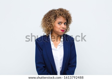 young woman african american with a goofy, crazy, surprised expression, puffing cheeks, feeling stuffed, fat and full of food against flat wall