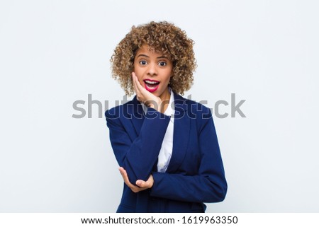 young woman african american open-mouthed in shock and disbelief, with hand on cheek and arm crossed, feeling stupefied and amazed against flat wall