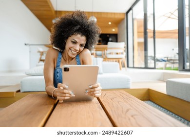 A young woman of African descent in a modern, sunlit living room, smiling as she uses a tablet. She has curly hair and is dressed casual, appearing relaxed and happy. Cozy and inviting atmosphere. - Powered by Shutterstock