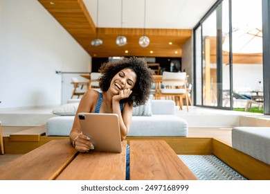 A young woman of African descent in a modern, sunlit living room, smiling as she uses a tablet. She has curly hair and is dressed casually, appearing relaxed and happy. The co and inviting atmosphere - Powered by Shutterstock