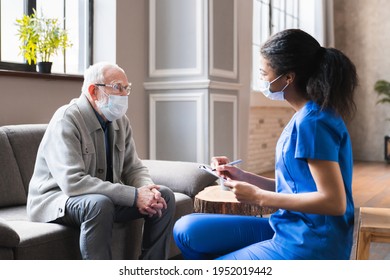 Young woman african doctor give a consultation to elderly old senior grandfather man during home visit in medical mask against Covid 19, nurse visit senior male patient at home consult 