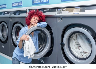 A Young Woman With Afro Hair Leaning Against The Door Of The Industrial Washing Machine Holding The Clothes In A Blue Automatic Laundromat